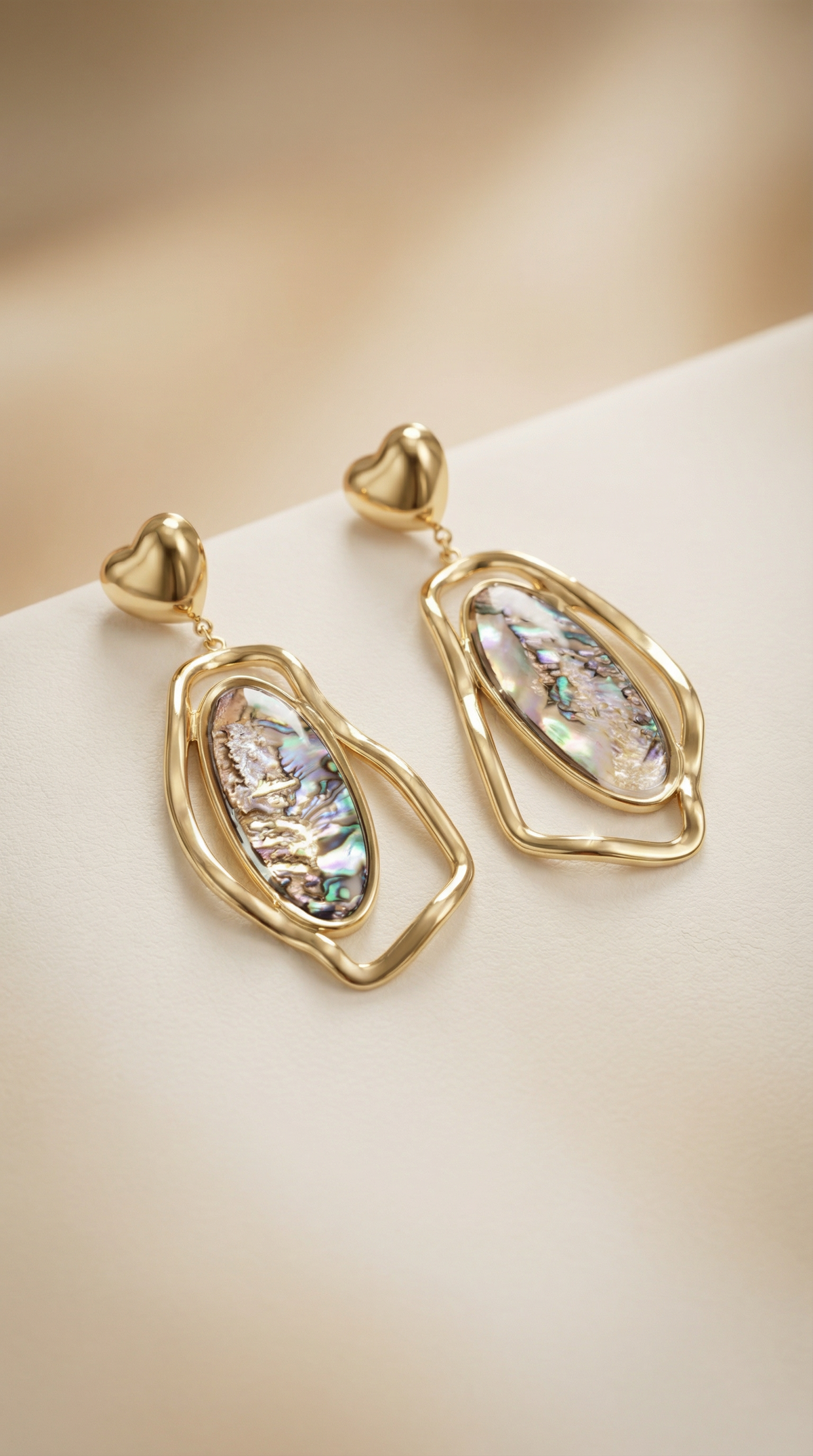 A pair of gold dangle earrings with heart-shaped posts and irregular gold frames around iridescent abalone shell inlays, displayed on a soft beige surface.