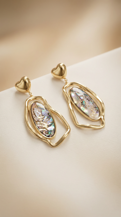 A pair of gold dangle earrings with heart-shaped posts and irregular gold frames around iridescent abalone shell inlays, displayed on a soft beige surface.